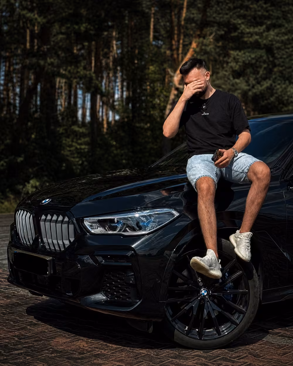 Руслан рядом с BMW в сосновом лесу в стилистике No Limits